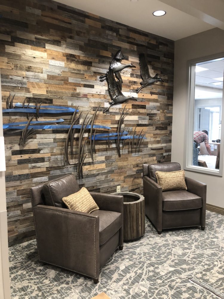 A trio of bronze sandhill cranes fly across a scene of tall prairie grasses and a rich blue river representing the Platte River. The piece is fixed to a mixed finished wood wall. At Vetter Health Services in Kearney Nebraska.
