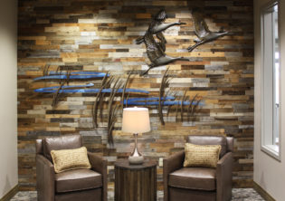 A trio of bronze sandhill cranes fly across a scene of tall prairie grasses and a rich blue river representing the Platte River. The piece is fixed to a mixed finished wood wall. At Vetter Health Services in Kearney Nebraska.