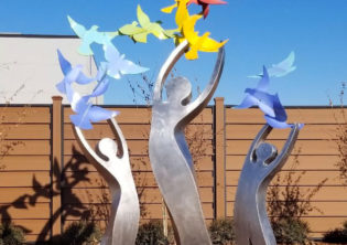 These three stainless steel figures represent a mother, and her two children, a boy and a girl. Their arms joyously reach upward as a rainbow of birds fly from their arms. Representing the endless possibilities that tomorrow brings. Located in