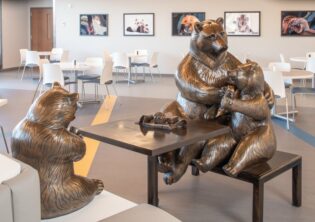 Mother bear with her two cubs, sitting at a table in the cafeteria eating lunch. One cub is eating out of a jar of honey, while the other is eating a paenut butter and jelly sandwich. The mama bear is claening the sandwich eating bear's face with a napkin. Bronze sculpture in Children's Hospital and Medical Center in Omaha, Nebraska.