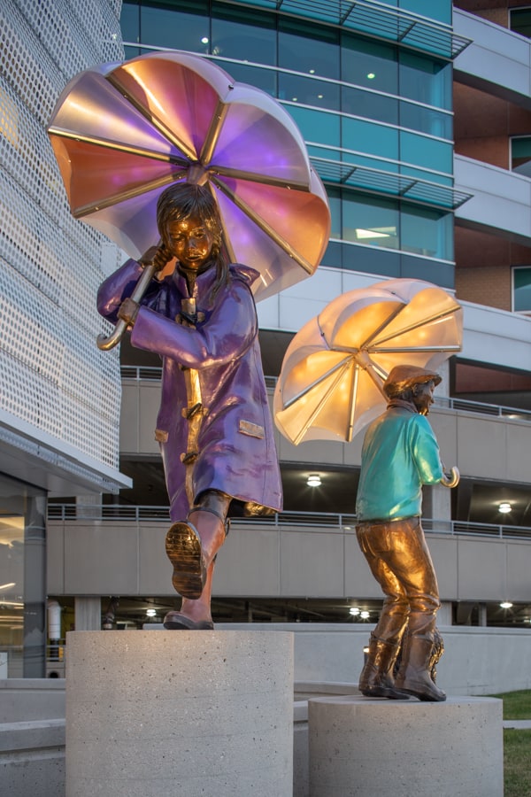 A young asain girl in a purple raincoat with orange rainboots is marching through the rain holding an umbrella standing next to the boy in the green sweater with shnauzer by his side. Boy in green sweater and driver's cap with a scarf, holds an umbrella. A miniature shnauzer sits patiently beside him. Imagine sculpture installation, Children's HOspital and Medical Center Omaha, Nebraska.