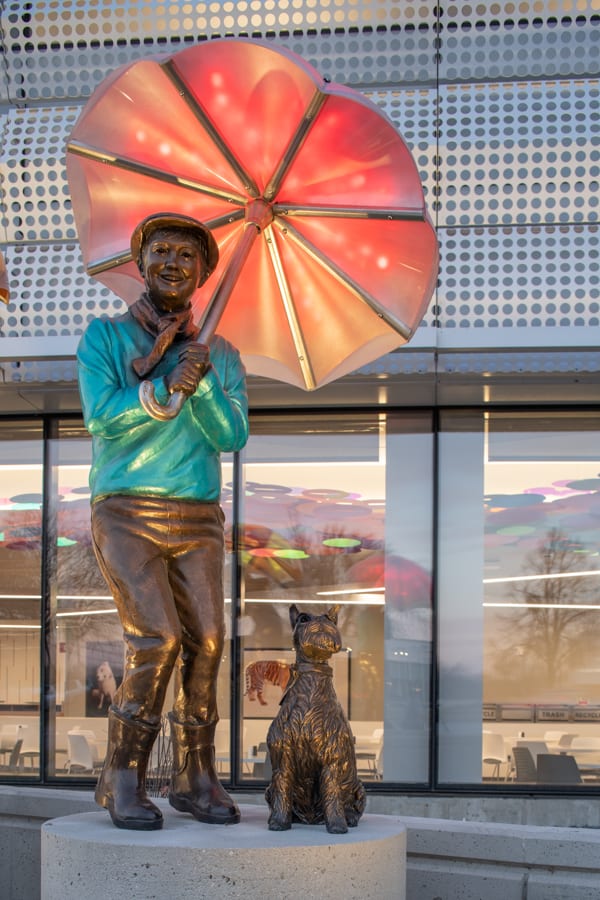 Boy in green sweater and driver's cap with a scarf, holds an umbrella. A miniature shnauzer sits patiently beside him. Imagine sculpture installation, Children's HOspital and Medical Center Omaha, Nebraska.