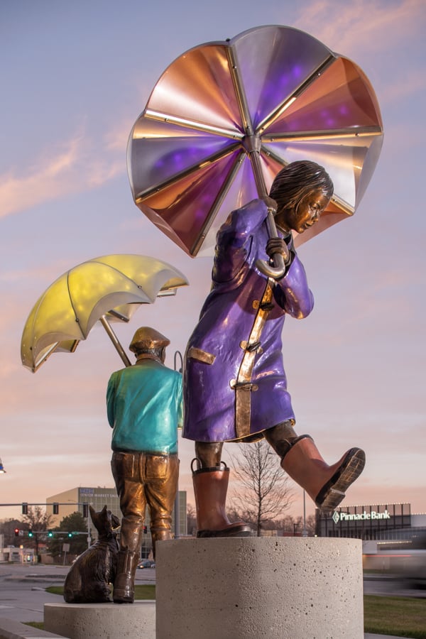 A young asain girl in a purple raincoat with orange rainboots is marching through the rain holding an umbrella standing next to the boy in the green sweater with shnauzer by his side. Boy in green sweater and driver's cap with a scarf, holds an umbrella. A miniature shnauzer sits patiently beside him. Imagine sculpture installation, Children's HOspital and Medical Center Omaha, Nebraska.