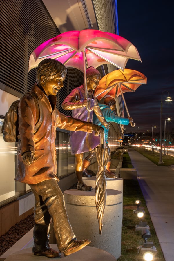 Three bronze children holding umbrellas. A small Boy in an orange raincoat holds a closed umbrella, the african american girl in the middle wearing a purple jumper holind an umbrella above her head. The boy on the end wearing a turquoise rain coat and orange rain boots is holding a copper umbrella. Imagine Children's Hospital and Medical Center in Omaha, Nebraska.